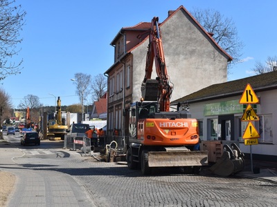 Na zdjęciu koparko-ładowarka, w tle prace budowlane w przestrzeni miejskiej.