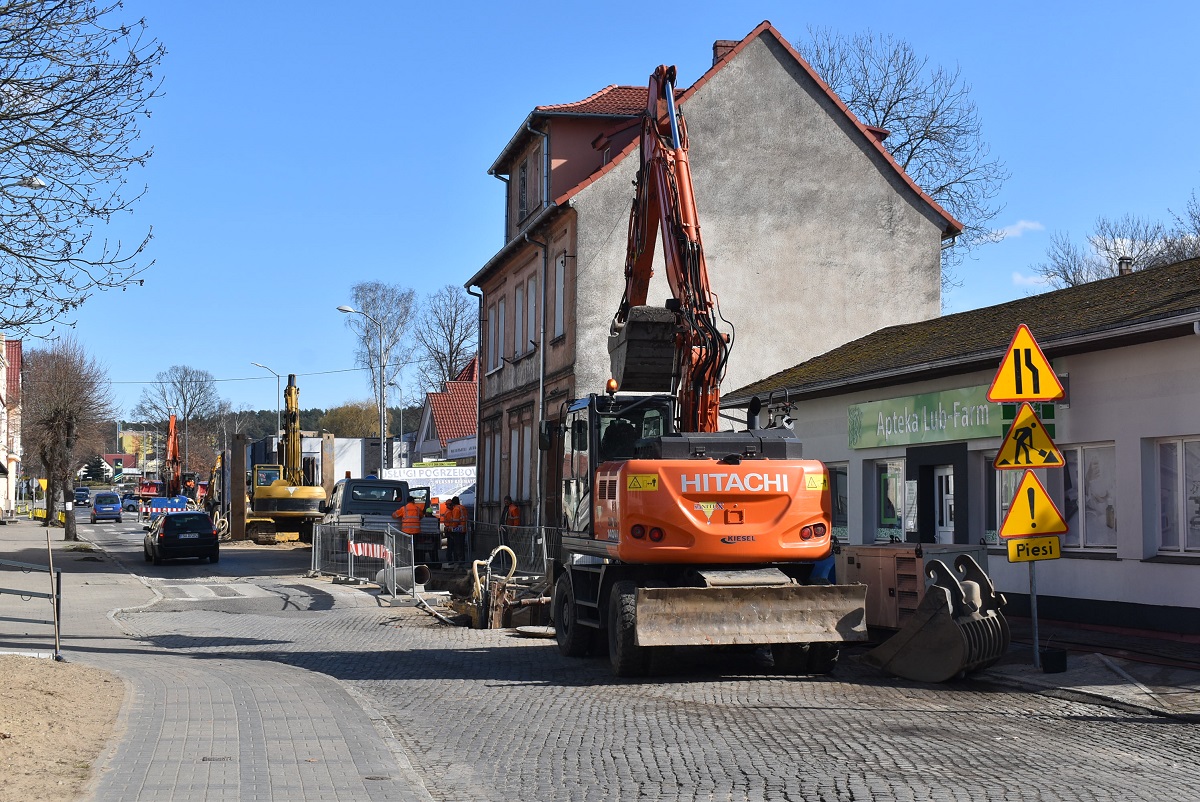 Na zdjęciu koparko-ładowarka, w tle prace budowlane w przestrzeni miejskiej.