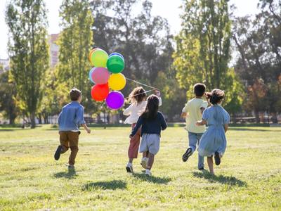 Zdjęcie przedstawia piątkę biegnących dzieci. Dziewczynka na przodzie trzyma balony.