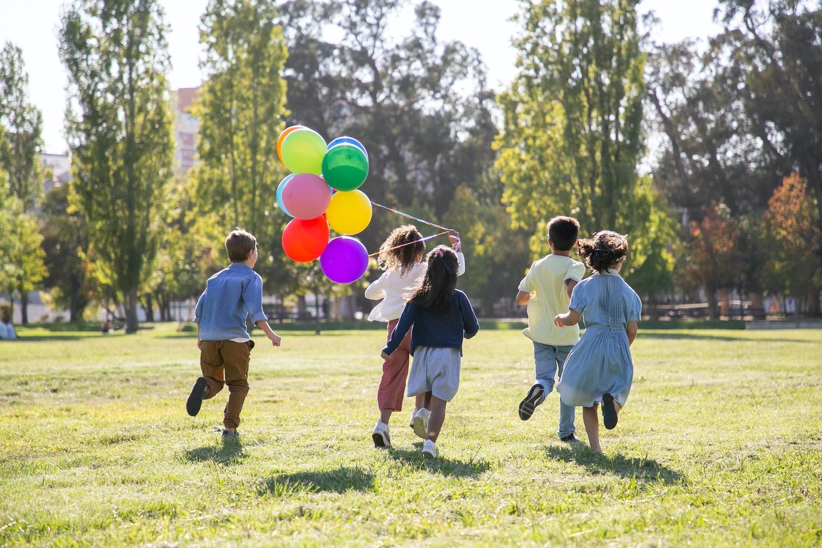 Zdjęcie przedstawia piątkę biegnących dzieci. Dziewczynka na przodzie trzyma balony.