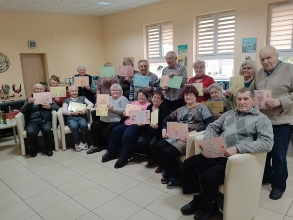 Alt="Zdjęcie przedstawia Seniorów z Domu Senior+, z laurkami przygotowanymi przez przedszkolaków z Ciasnej."