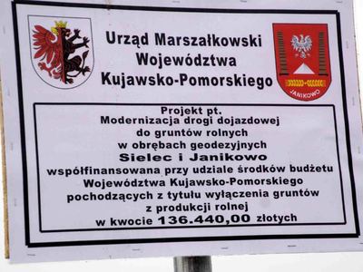 Zdjęcie białej tablicy informacyjnej z czarnymi napisami i 2 kolorowymi logami na której znajduje kolorowe Logo Urzędu Marszałkowskiego Województwa Kujawsko Pomorskiego i Logo Gminy Janikowo oraz informacja Projekt pt Modernizacja drogi dojazdowej do gruntów rolnych w obrębach geodezyjnych Janikowo i Sielec współfinansowana przy udziale środków budżetu Województwa Kujawsko Pomorskiego pochodzących z tytułu wyłączenia gruntów z produkcji rolnej w kwocie 136 44000 złotych [400x300]