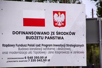 Tablica biała z flagą biało czerwoną oraz logiem Polski Ład. Na tablicy czarnym tekstem wymieniona jest nazwa inwestycji oraz forma i wysokość dofinansowania.