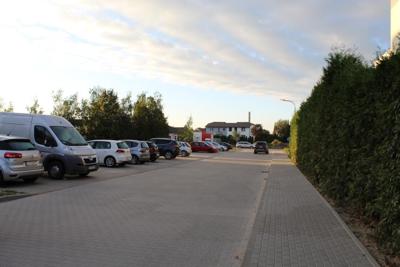 Zdjęcie przedstawia ulicę z szarej kostki brukowej. Po lewej stronie prostopadle do ulicy parkują samochody. Po prawej biegnie chodnik. Za chodnikiem widoczny jest żywopłot.