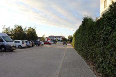 Zdjęcie przedstawia ulicę z szarej kostki brukowej. Po lewej stronie prostopadle do ulicy parkują samochody. Po prawej biegnie chodnik. Za chodnikiem widoczny jest żywopłot.