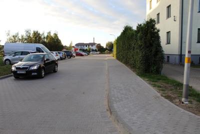 Zdjęcie przedstawia ulicę z szarej kostki brukowej. Po lewej stronie prostopadle do ulicy parkują samochody. Po prawej biegnie chodnik. Za chodnikiem widoczny jest żywopłot. Za żywopłotem widać część bloko mieszkalnego.