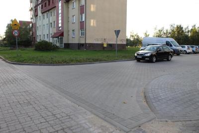 Zdjęcie pokazuje wjazd na ulicę z szarej kostki brukowej. Za wjazdem jest blok mieszkalny. Na prawej stronie zdjęcia parkują samochody.