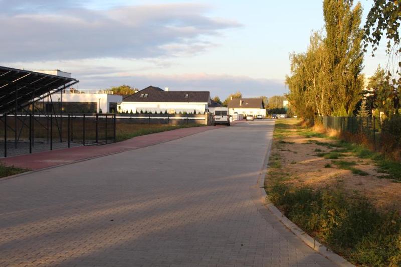 Zdjęcie przedstawia ulicę z szarej kostki brukowej. Na lewej stronie znajduje się pas z czerwonej kostki brukowej. Na czerwonej części parkują trzy samochody. W oddali widać domy jednorodzinne.