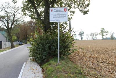 Na zdjęciu znajsuje się sroga asfaltowa w kolorze ciemnym z poboczem w Kororze jasnym. Przy drodze widać pas gruntu z zieloną trawą oddzielającą drogę od pola uprawnego na którym widać resztki uprawy kukurydzy po zbiorze. Na pasie trawnika ustawiona tablica informacyjna na tle drzewa i częściowo nieba. Na tablicy znajdują się dla loga. Województwa Kujawsko Pomorskiego oraz Gminy Janikowo. Napis w kolorze czarnym mówiący: Urząd Marszałkowski Województwa Kujawsko Pomorskiego. Projekt pt. Modernizacja drogi dojazdowej do gruntów rolnych w obrębie geodezyjnym Sosnówiec o nawierzchni asfaltowej zrealizowanej w roku 2024, wspófinansowana przy udziale środków budżetu województwa Kujawsko Pomorskiego w kwocie 350 000,00 zł. Wartość dotacji
