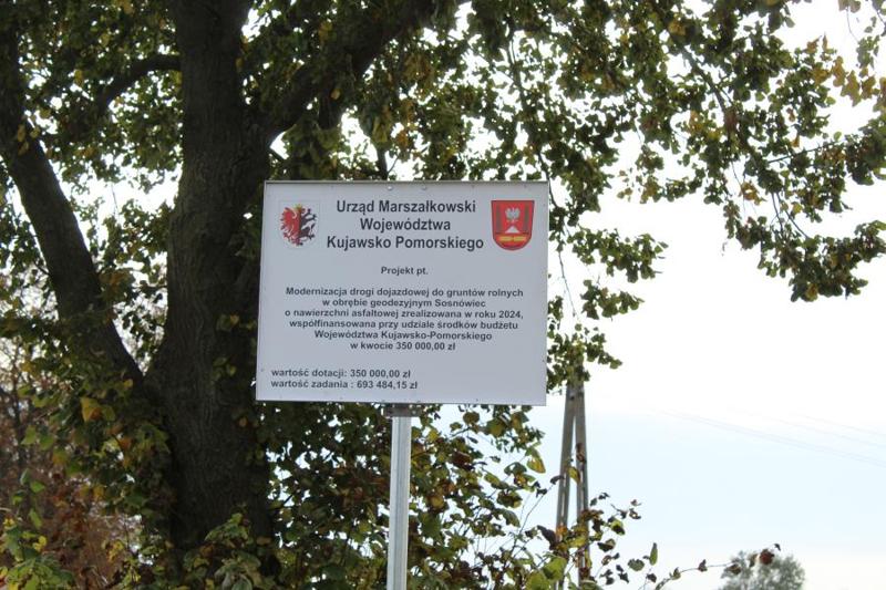 Na zdjęciu znajduje się biała tablica informacyjna na tle drzewa i częściowo nieba. Na tablicy znajdują się dwa loga. Województwa Kujawsko Pomorskiego oraz Gminy Janikowo. Napis w kolorze czarnym mówiący:  Urząd Marszałkowski Województwa Kujawsko Pomorskiego. Projekt pt. Modernizacja drogi dojazdowej do gruntów rolnych w obrębie geodezyjnym Sosnówiec o nawierzchni asfaltowej zrealizowanej w roku 2024, wspófinansowana przy udziale środków budżetu województwa Kujawsko Pomorskiego w kwocie 350 000,00 zł. Wartość dotacji 350 000,00zł. Wartość zadania 693 484,15 zł