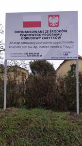 Zdjęcie białej tablicy z Flagą oraz Godłem Polski. Na tablicy znajdują się napisy w kolorze czarnym opisujące poziom dofinansowania oraz tytuł zadania. W tle widać fragmenty budynków oraz zieleni (trawy i krzewów).
