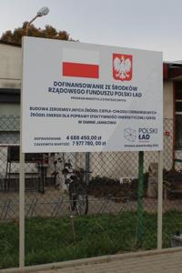 Tablice białe zawierające znak flagi polski biało czerwonej oraz godło polski. Czarnymi literami napisany jest tytuł inwestycji BUDOWA ZEROEMISYJNYCH ŹRÓDEŁ CIEPŁA-CHŁODU I ODNAWIALNYCH ŹRÓDEŁ ENERGII DLA POPRAWY EFEKTYWNOŚCI ENERGETYCZNEJ SZKÓŁ W GMINIE JANIKOWO oraz wartości dofinansowania i wartość inwestycji.