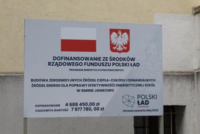 Tablice białe zawierające znak flagi polski biało czerwonej oraz godło polski. Czarnymi literami napisany jest tytuł inwestycji BUDOWA ZEROEMISYJNYCH ŹRÓDEŁ CIEPŁA-CHŁODU I ODNAWIALNYCH ŹRÓDEŁ ENERGII DLA POPRAWY EFEKTYWNOŚCI ENERGETYCZNEJ SZKÓŁ W GMINIE JANIKOWO oraz wartości dofinansowania i wartość inwestycji.