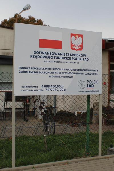 Tablice białe zawierające znak flagi polski biało czerwonej oraz godło polski. Czarnymi literami napisany jest tytuł inwestycji BUDOWA ZEROEMISYJNYCH ŹRÓDEŁ CIEPŁA-CHŁODU I ODNAWIALNYCH ŹRÓDEŁ ENERGII DLA POPRAWY EFEKTYWNOŚCI ENERGETYCZNEJ SZKÓŁ W GMINIE JANIKOWO oraz wartości dofinansowania i wartość inwestycji.
