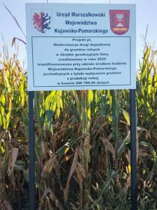 Rosnącą kukurydza. Przed kukurydzą stoi biała tablica, na której są loga Województwa i gminy Janikowo oraz czarny tekst. Treść tekstu: Urząd Marszałkowski Województwa Kujawsko-Pomorskiego. W ramce znajduje się kolejny tekst: Projekt pn. „Modernizacja drogi dojazdowej do gruntów rolnych w obrębie geodezyjnym Góry zrealizowana w roku 2025, współfinansowana przy udziale środków budżetu Województwa Kujawsko -Pomorskiego w kwocie 260 780,00 zł”.
