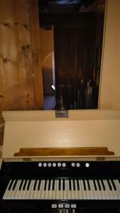 Pomieszczenie w którym znajduje sie fragment organów z klawiszami w kolorach biało czarnych. widać podniesiona pokrywę w kolorze cielistym z elementem drewna w kolorze brązowym zabezpieczającą klawiaturę. W tle elementy rurowe piszczałem  oraz po lewej stronie fragment ściany drewnianej w kolorze jasnobrązowym z widocznymi słojami drewna. centralnie ciemnobrązowy element w kształcie prostokąta o nieokreślonej funkcji.