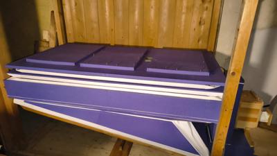 Elementy miecha wtłaczającego powietrze niezbędne do wydawania dźwięków przez piszczałki. element jest w kolorach fioletowo białych ułożonych w poziomie naprzemiennie. całość osadzona na drewnianej ramie w kolorze jasnego lakierowanego drewna. za miechem widać fragment ściany z desek w kolorze pomarańczowo jasnobrązowym.