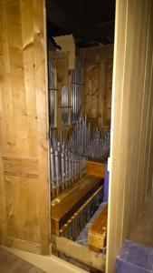 Pomieszczenie z jasnego drewna w którego wnętrzu widoczne są ustawione pionowo srebrne rury (piszczałki) z obwódkami w górnej części w kolorze czerwono- brązowym. Rury są poustawiane w rzędach na różnej wysokości. Rury są różnej długości i średnicy w dolnej czści widoczne są przewody w kolorze ciemnym kilkadziesiąt szt. sufit pomieszczenia w ciemnym kolorze.