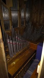 Pomieszczenie z jasnego drewna w którego wnętrzu widoczne są ustawione pionowo srebrne rury (piszczałki) z obwódkami w górnej części w kolorze czerwono- brązowym. Rury są poustawiane w rzędach na różnej wysokości. Rury są różnej długości i średnicy w dolnej czści widoczne są przewody w kolorze ciemnym kilkadziesiąt szt. sufit pomieszczenia w ciemnym kolorze.