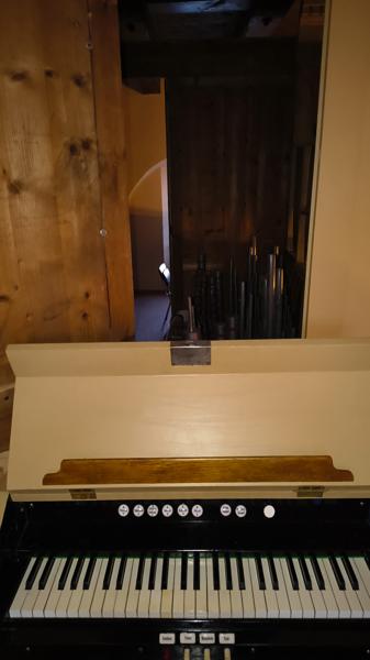 Pomieszczenie w którym znajduje sie fragment organów z klawiszami w kolorach biało czarnych. widać podniesiona pokrywę w kolorze cielistym z elementem drewna w kolorze brązowym zabezpieczającą klawiaturę. W tle elementy rurowe piszczałem  oraz po lewej stronie fragment ściany drewnianej w kolorze jasnobrązowym z widocznymi słojami drewna. centralnie ciemnobrązowy element w kształcie prostokąta o nieokreślonej funkcji.