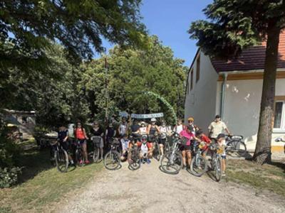 Grupa stojących rowerzystów. Po prawej szczyt domu, po lewej mały budynek. W tle drzea.