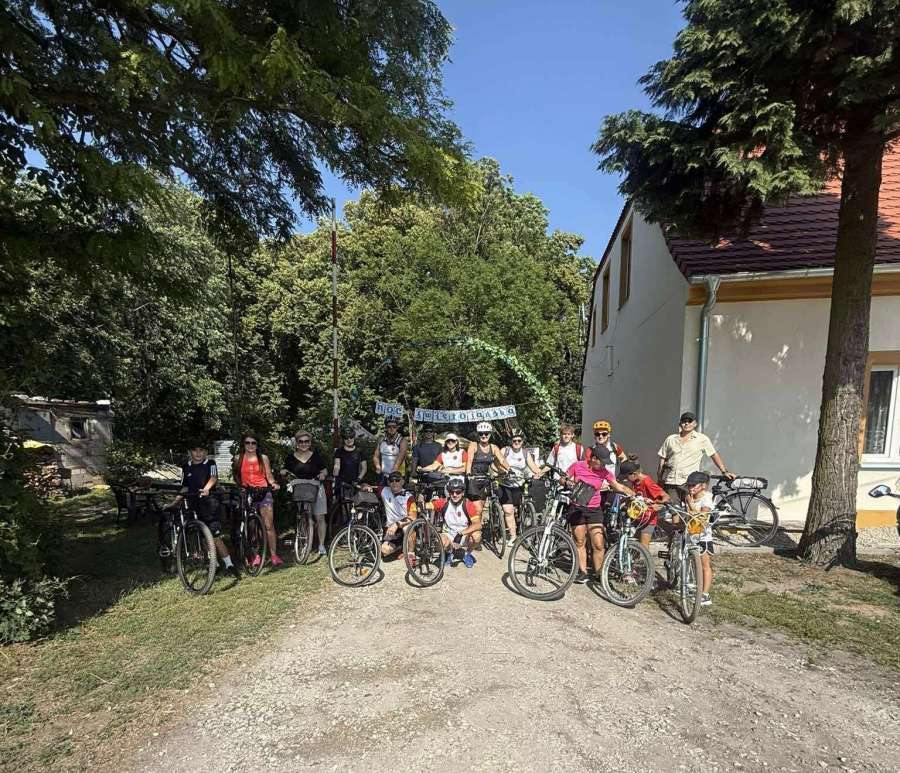 Grupa stojących rowerzystów. Po prawej szczyt domu, po lewej mały budynek. W tle drzea.