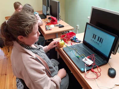 Uczennica siedzi przy biurko. Na biurku leży uruchomiony laptop z wyświetlonym fragmentem klawiatury pianina. Na biurku leżą kolorowe kable: czerwony, zielony żółty i biały. Czerwony kabel podłączony jest do komputera, a końcówka zielonego wbita jest w jabłko leżące na lewo od laptopa. Uczennica dotyka palcami lewej ręki jabłka. Na drugim planie widać podobne stanowisko, przy którym siedzą dwie uczennice.