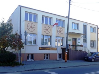 Dwupoziomowy budynek pomalowany na jasny żółty kolor z brązowym fundamentem. Do budynku prowadzą betonowe schody z kwiatami na poręczy z metalu. Między trzema oknami pierwszego i drugiego poziomu namalowane koła ze szprychami. Na ścianie pomarańczowy napis na brązowym tle: Bursztynowy Senior.