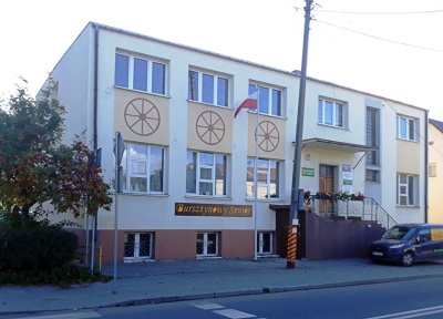 Dwupoziomowy budynek pomalowany na jasny żółty kolor z brązowym fundamentem. Do budynku prowadzą betonowe schody z kwiatami na poręczy z metalu. Między trzema oknami pierwszego i drugiego poziomu namalowane koła ze szprychami. Na ścianie pomarańczowy napis na brązowym tle: Bursztynowy Senior.