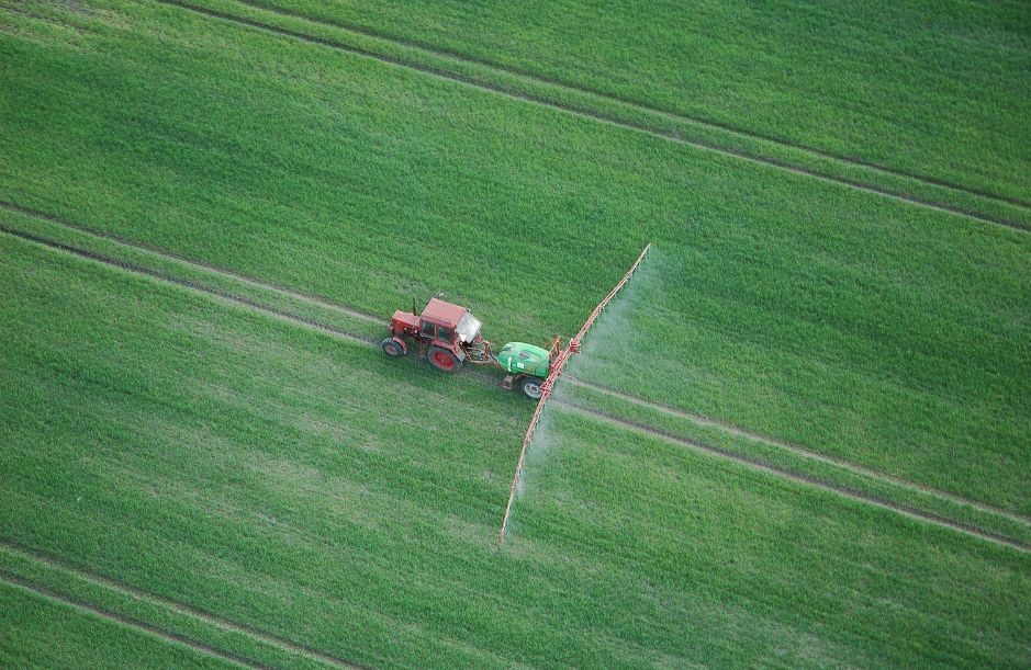 Zdjęcie przedstawia traktor i opryskiwacz jadące po zielonym polu uprawnym