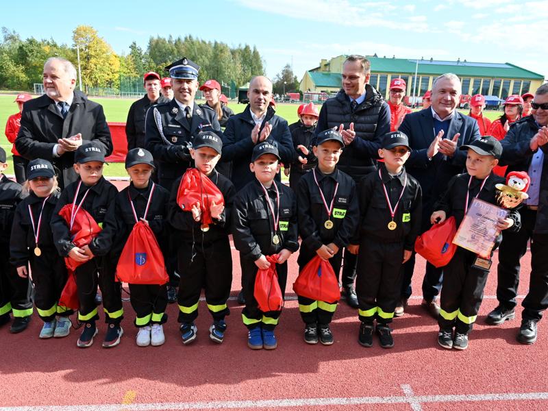 Grupa dzieci w strojach strażackich z medalami oraz mężczyźni, w tym przedstawiciel straży pożarnej, stoją podczas uroczystości na bieżni sportowej.