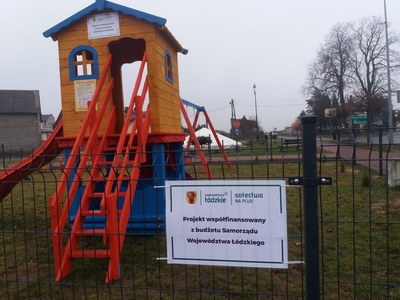 Na pierwszym planie płot z tabliczką o treści Projekt współfinansowany z budżetu Samorządu Województwa Łódzkiego, w tle zjeżdżalnia w formie domku.