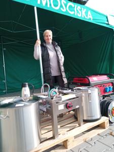 Sołtys wsi Mościska stojąca pod namiotem, na pierwszym planie: agregat prądotwórczy, przedłużacz, taboret gastronomiczny, zestaw naczyń plenerowych