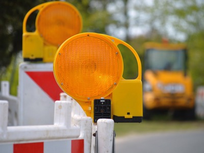 Na pierwszym planie zapora drogowa i ostrzegawcza lampa drogowa, w tle urządzenie budowlane