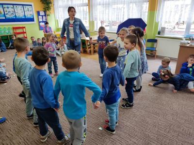 Grupa dzieci wraz z panią bawiąca się w kółku. Wewnątrz.
