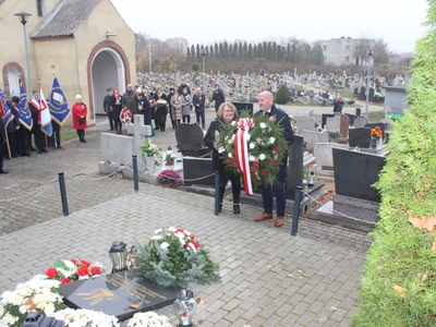 Zdjęcie wykonane na zewnątrz. Na zdjęciu widoczne osoby składające wiązankę kwiatów na grobie poległych w obronie Ojczyzny.