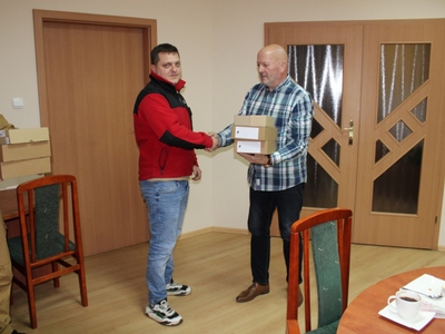 Wewnątrz. Dwie osoby stoją. Jedna drugiej wręcza paczkę.