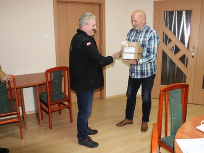 Wewnątrz. Dwie osoby stoją. Jedna drugiej wręcza paczkę.