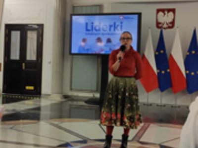 Zdjęcie wykonane wewnątrz pomieszczenia. Na zdjęciu widoczna kobieta. W tle godło RP oraz flagi RP i UE.