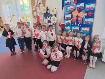 Grupa dzieci pozująca do zdjęcia z przypiętymi kotylionami i flagami w narodowych barwach. Wewnątrz.