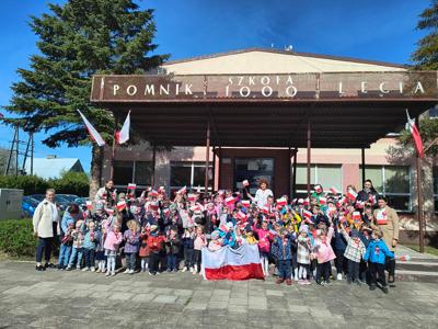 Grupa dzieci i nauczyciele pozujący do zdjęcia z flagami biało-czerwonymi. Na zewnątrz.
