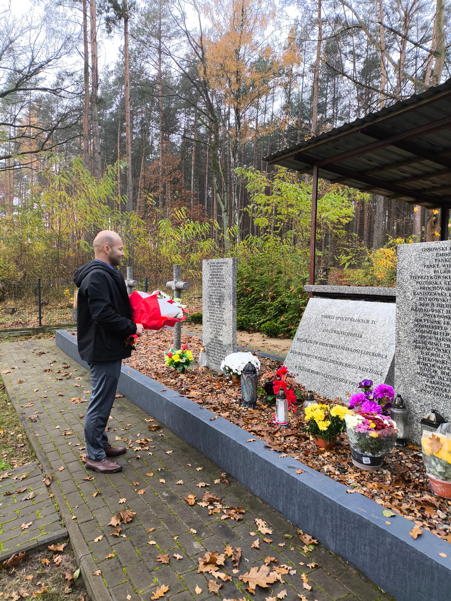 Zdjęcie przedstawia mogiłę zbiorową ogrodzoną płotem usytuowaną w lesie