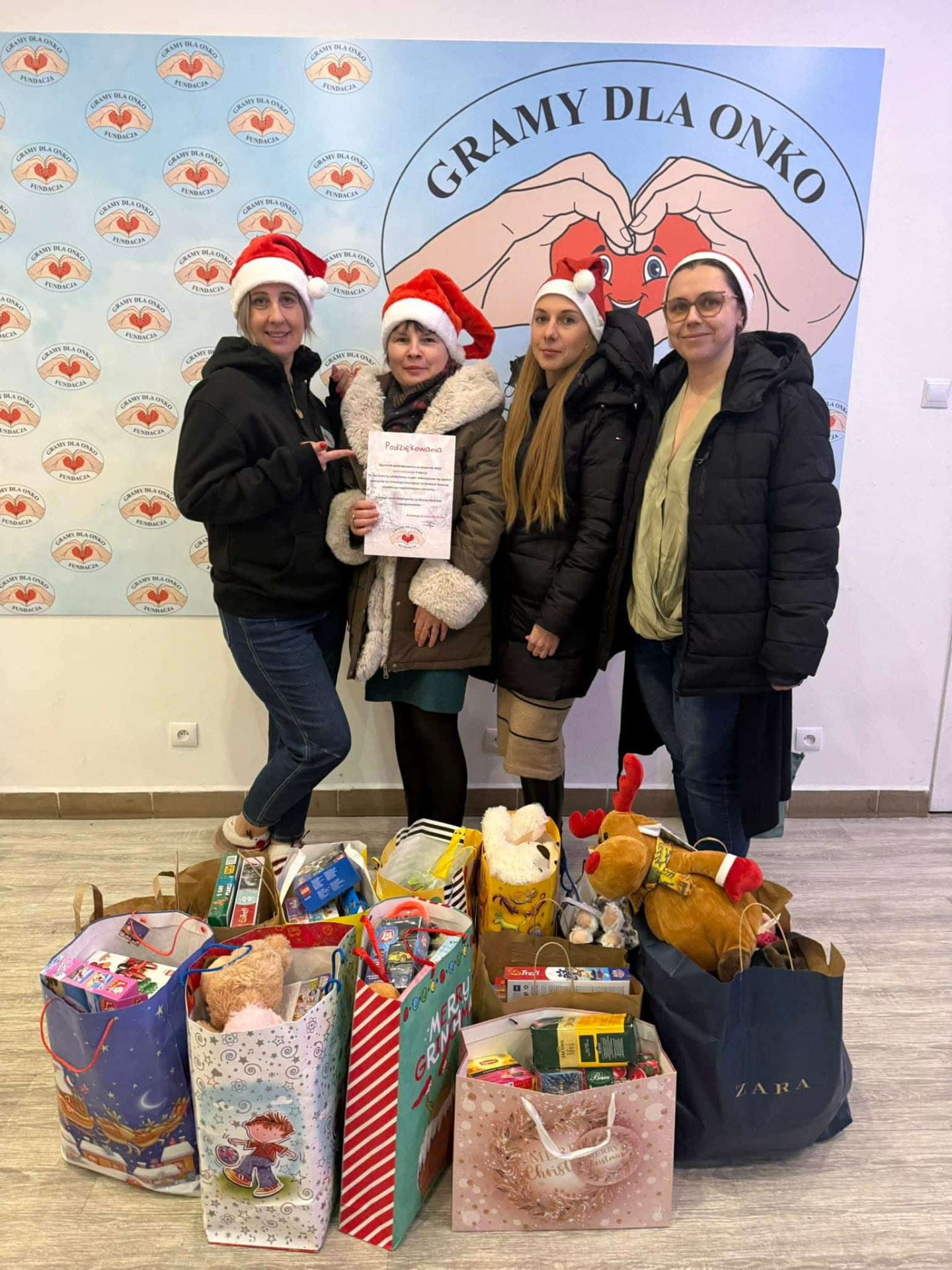 Cztery kobiety w czapkach Mikołaja pozujące do zdjęcia przy torbach z prezentami. Wewnątrz.