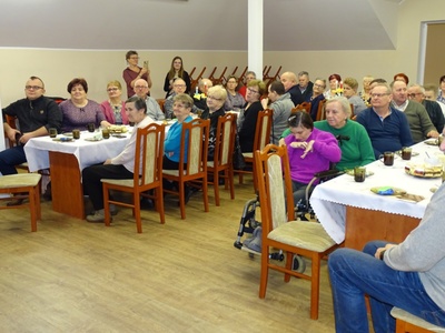 Zdjęcie przedstawia siedzących przy stole gości, którzy częstując się ciastem oglądają program artystyczny.