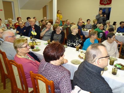 Zdjęcie przedstawia kolejnych siedzących przy stole gości, którzy częstując się oglądają program artystyczny.