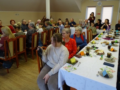 Grupa osób siedząca przy zastawionym stole  i przyglądająca się występom dzieci. Wewnątrz.