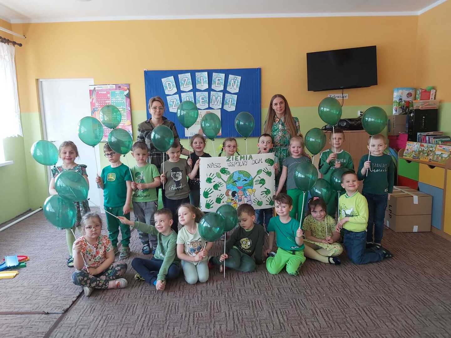 Grupa przedszkolaków z dwiema paniami opiekunkami ubrana na zielono i trzymająca baloniki. Wewnątrz.