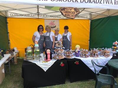 Zdjęcie wykonane wew. Namiotu. Na tle żółtej ściany widoczne cztery osoby reprezentujące koło KGW. Na stole przed nimi liczne smakołyki.