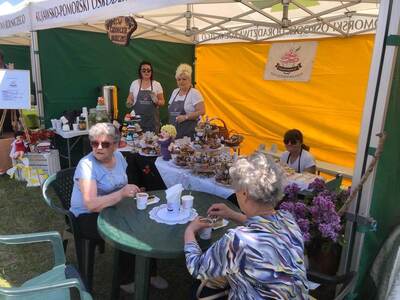 Dwie panie siedzące przy okrągłym stoliku spożywające kawę i słodki dodatek. W tle dwie osoby obsługujące stanowisko.