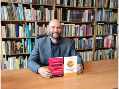 Autor Jacek Łapiński siedzący przy stole w tle regał z książkami.